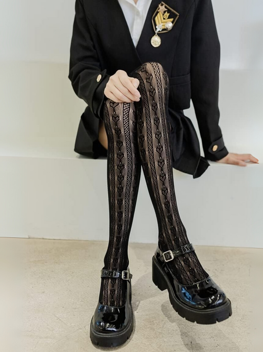 Person wearing a black blazer with a brooch, white shirt, black patterned stockings, and black shoes on a plain background.