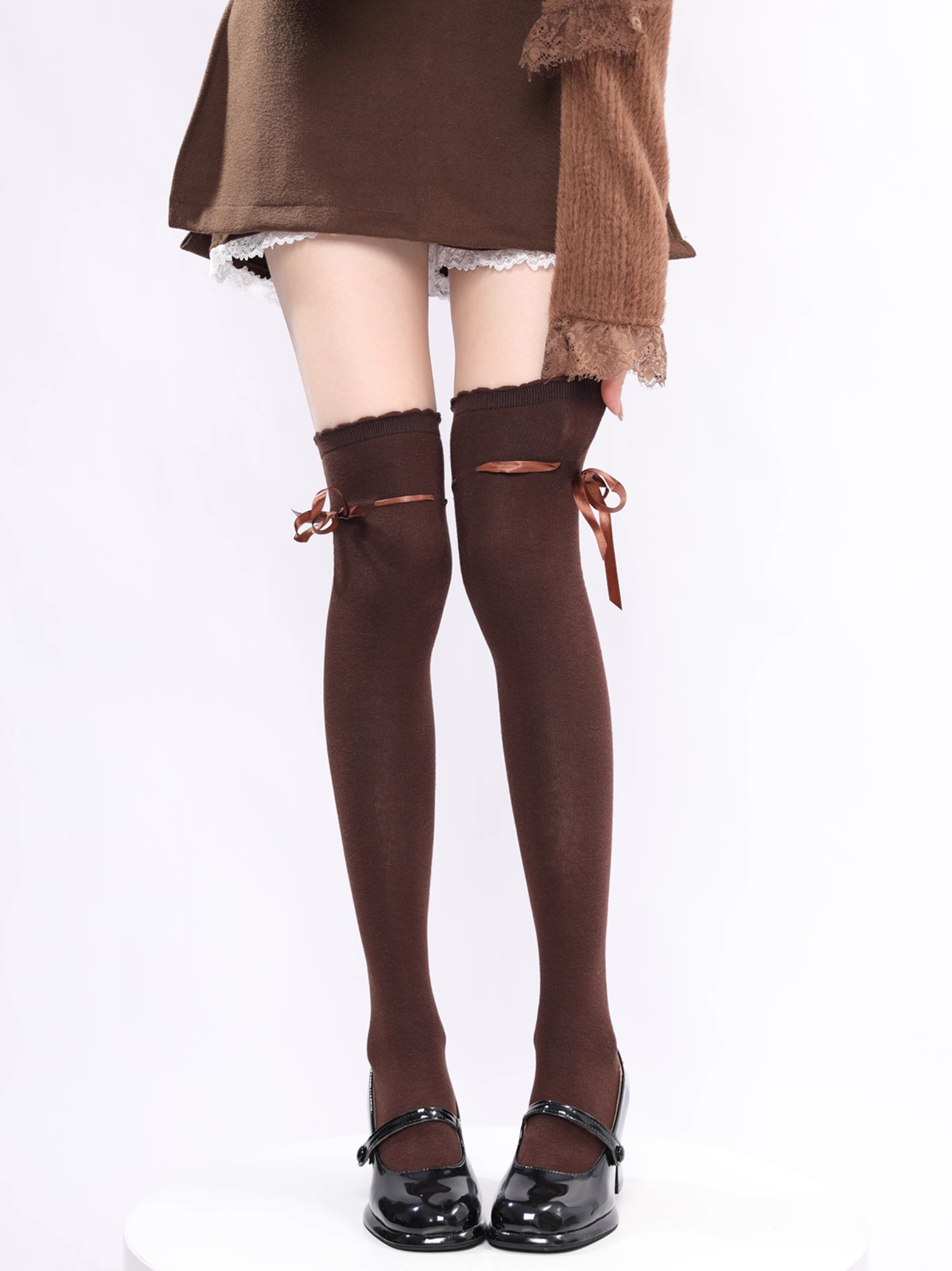 Brown knee-high socks with bow details worn over a white skirt and brown sweater on a white background.
